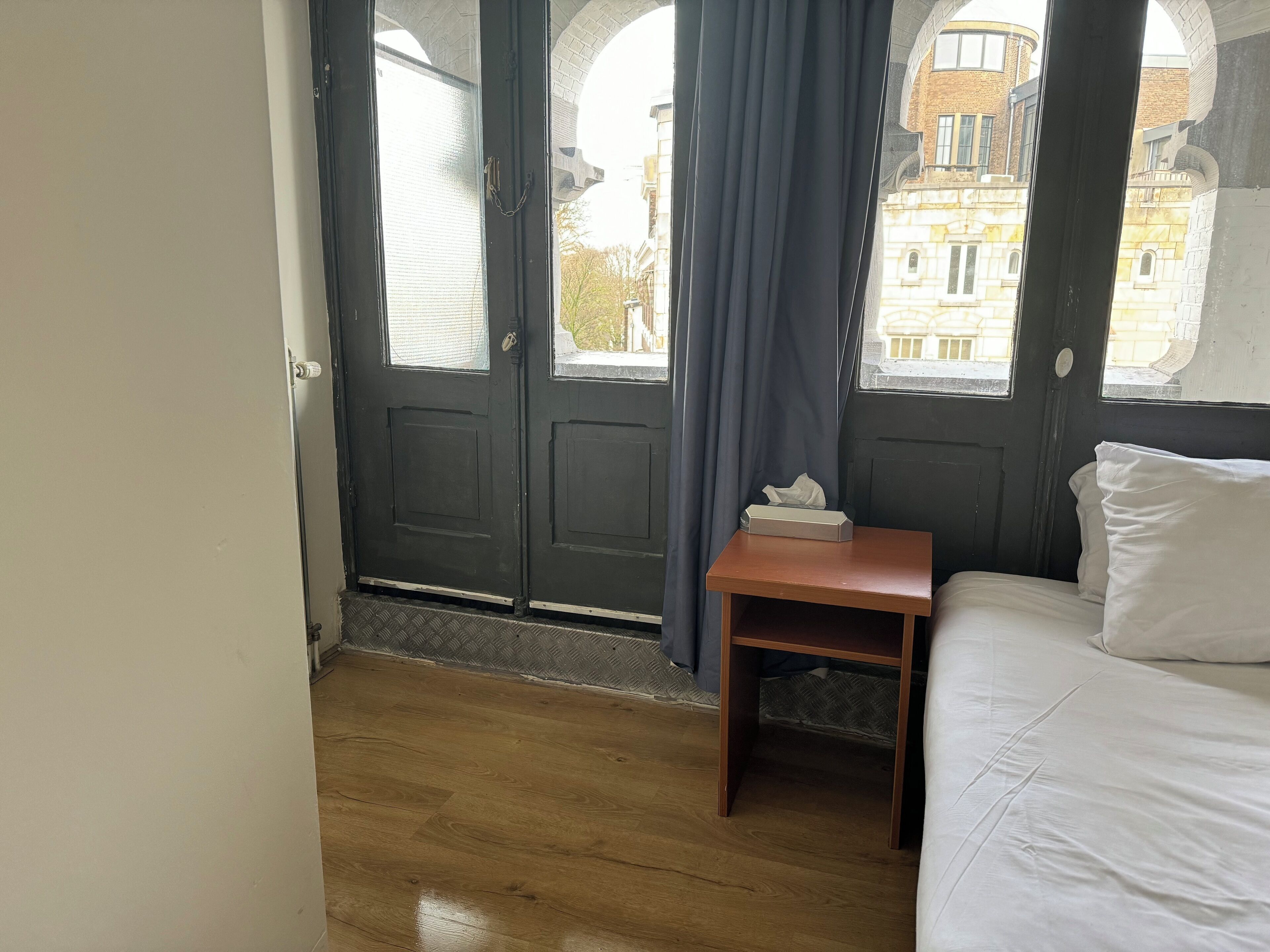 A room with a wooden floor, a bed with white bedding, a small wooden nightstand, a tissue box, and a view of a building through a dark door with a window.