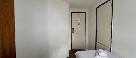 A hotel room with a wooden door, a white bed with a folded towel, a white wall, and a door with a no-smoking sign.