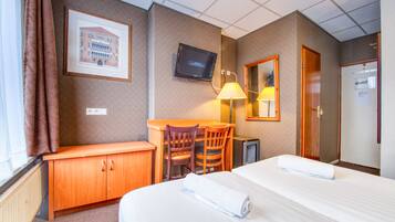 A hotel room with a bed, a desk with a chair, a television, a lamp, and a door.