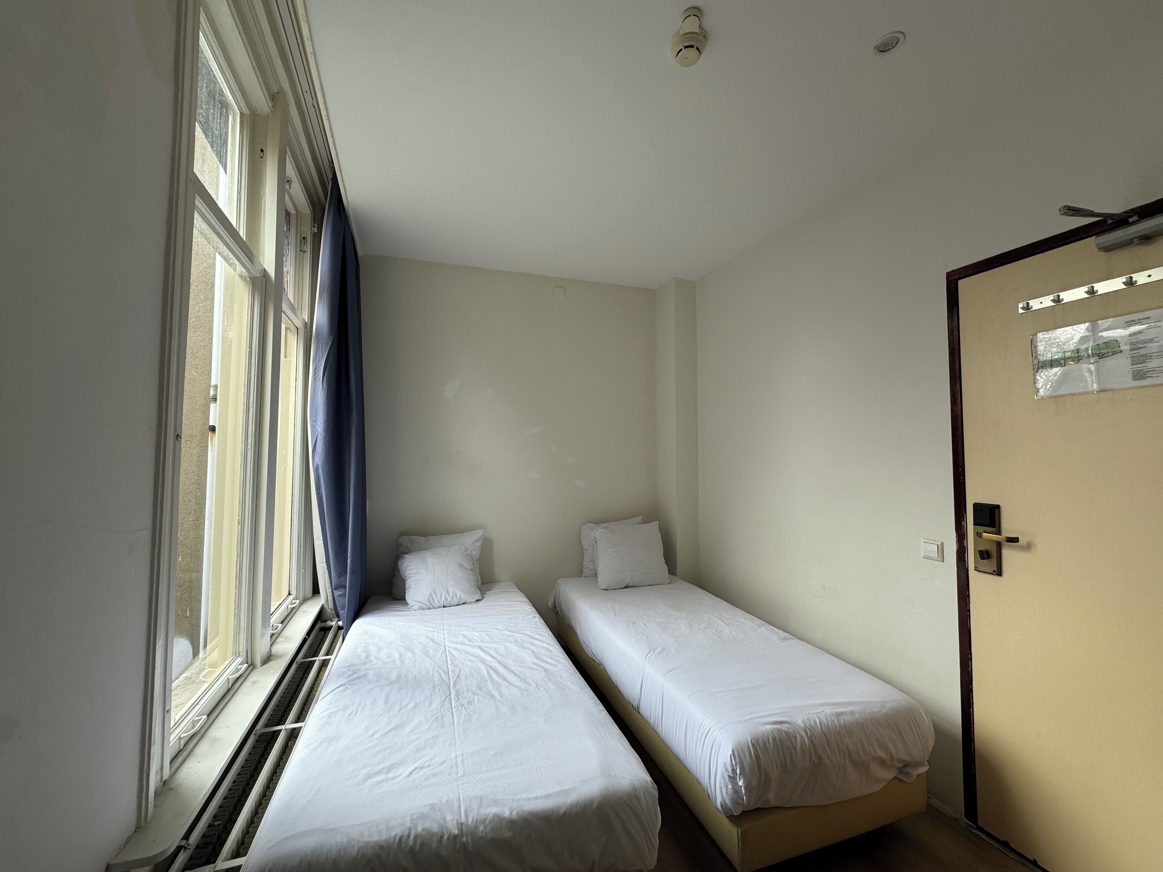 A small, single-room hotel room with two single beds, a window with curtains, a door with a sign, and a ceiling light.
