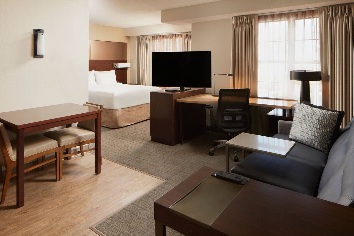 A hotel room with a bed, a desk with a computer, a TV, a sofa, and a dining table with chairs.