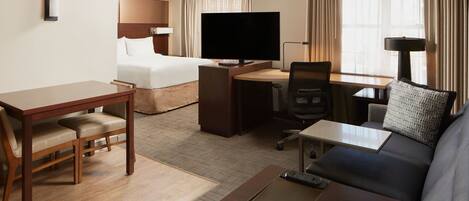 A hotel room with a bed, a desk with a computer, a TV, a sofa, and a dining table with chairs.
