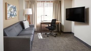 A hotel room with a gray sofa, a desk with a chair, a flat-screen TV, a lamp, and a window with curtains.