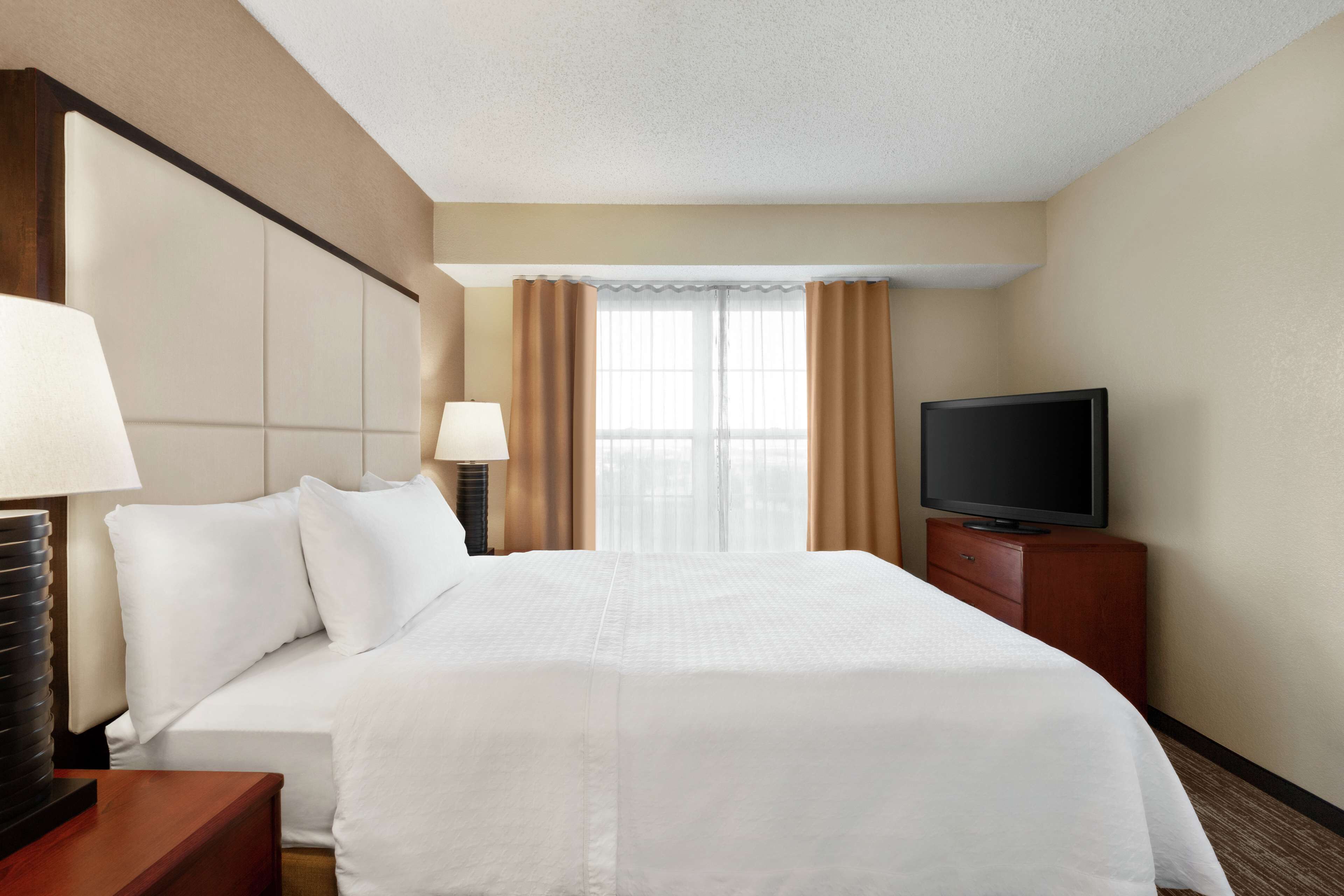 A hotel room with a large bed, a TV on a wooden stand, a nightstand, a lamp, and a window with curtains.