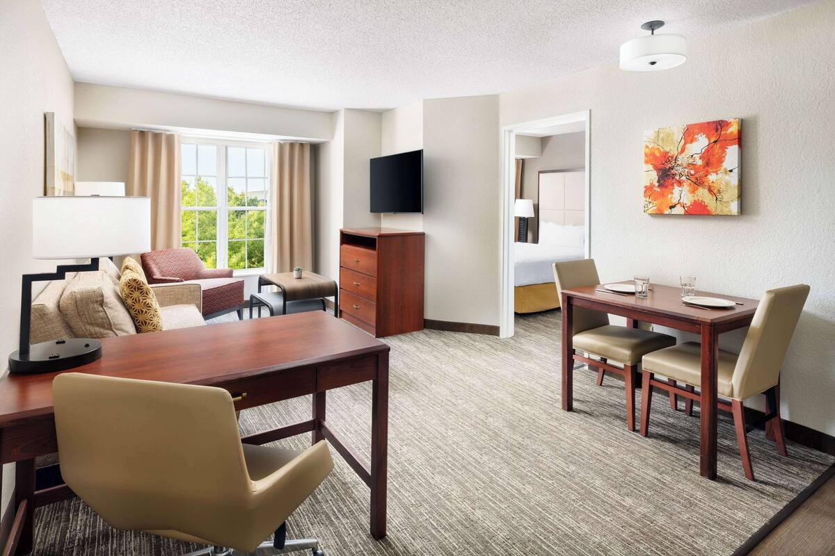 A hotel room with a dining area, a TV mounted on the wall, and a window with curtains.