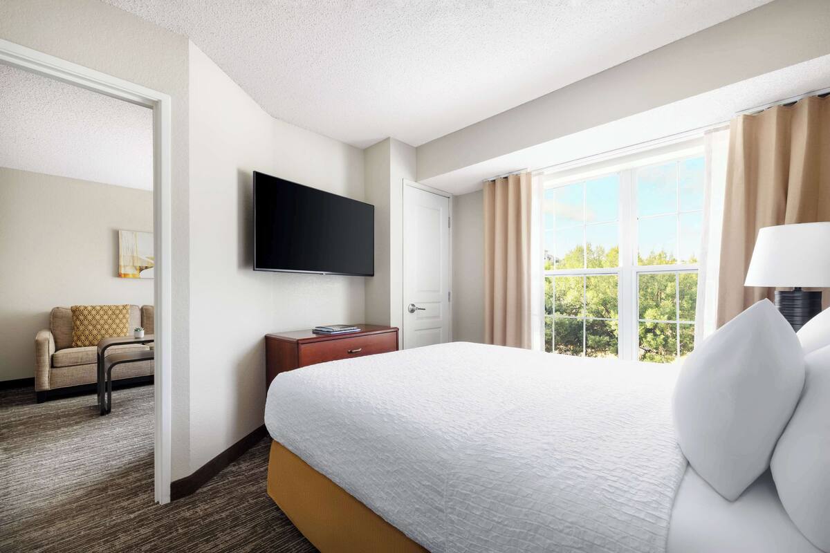 A hotel room with a large bed, a flat-screen TV mounted on the wall, a window with curtains, and a small seating area with a sofa and a table.