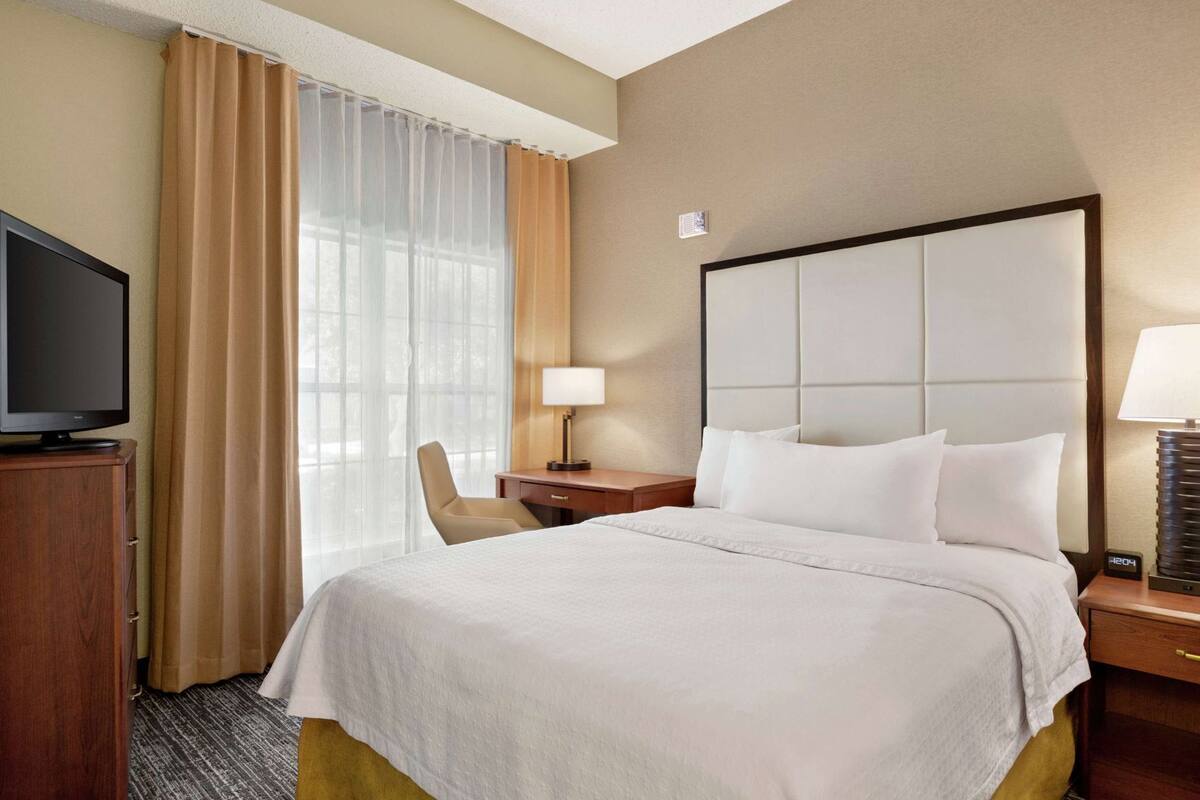 A hotel room with a bed, a TV, a desk, a chair, a lamp, and a clock.