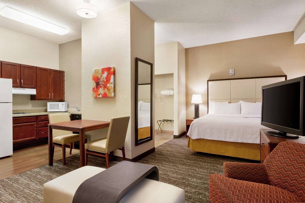 A hotel room with a bed, TV, desk, chair, microwave, and a painting on the wall.