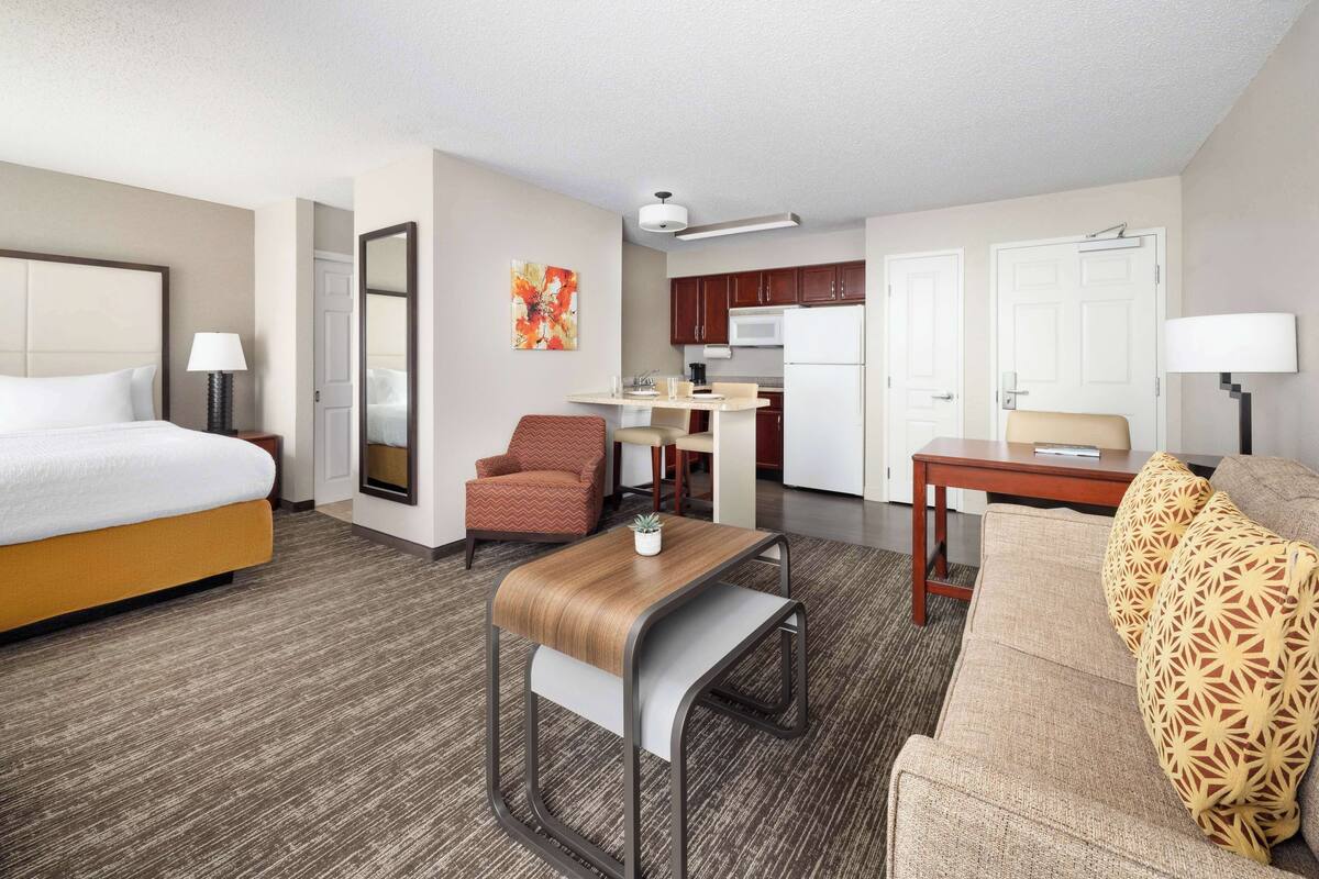 A hotel room with a bed, a sofa, a small table, a chair, a kitchenette, and a bathroom.