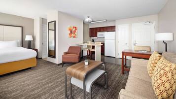A hotel room with a bed, a sofa, a small table, a chair, a kitchenette, and a bathroom.