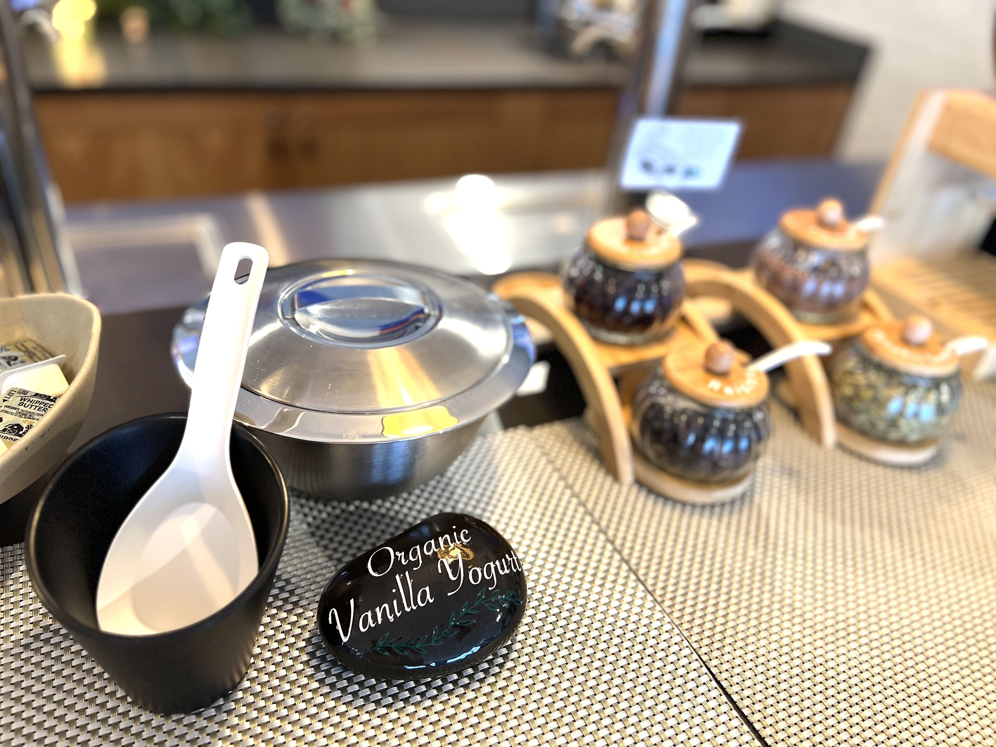 Uma bancada de cozinha com uma panela de aço inoxidável, uma tigela preta, uma colher e um prato com a etiqueta 'Organic Vanilla Yogurt'. Também há pequenos recipientes de cerâmica com tampas.