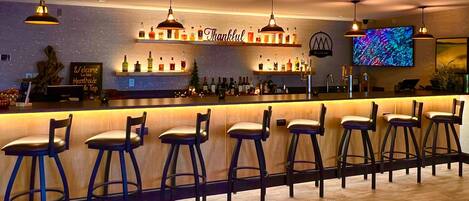 Eine Bar mit mehreren Barhocker, einer Theke mit einem 'Thankful'-Schild und einem Fernsehbildschirm.