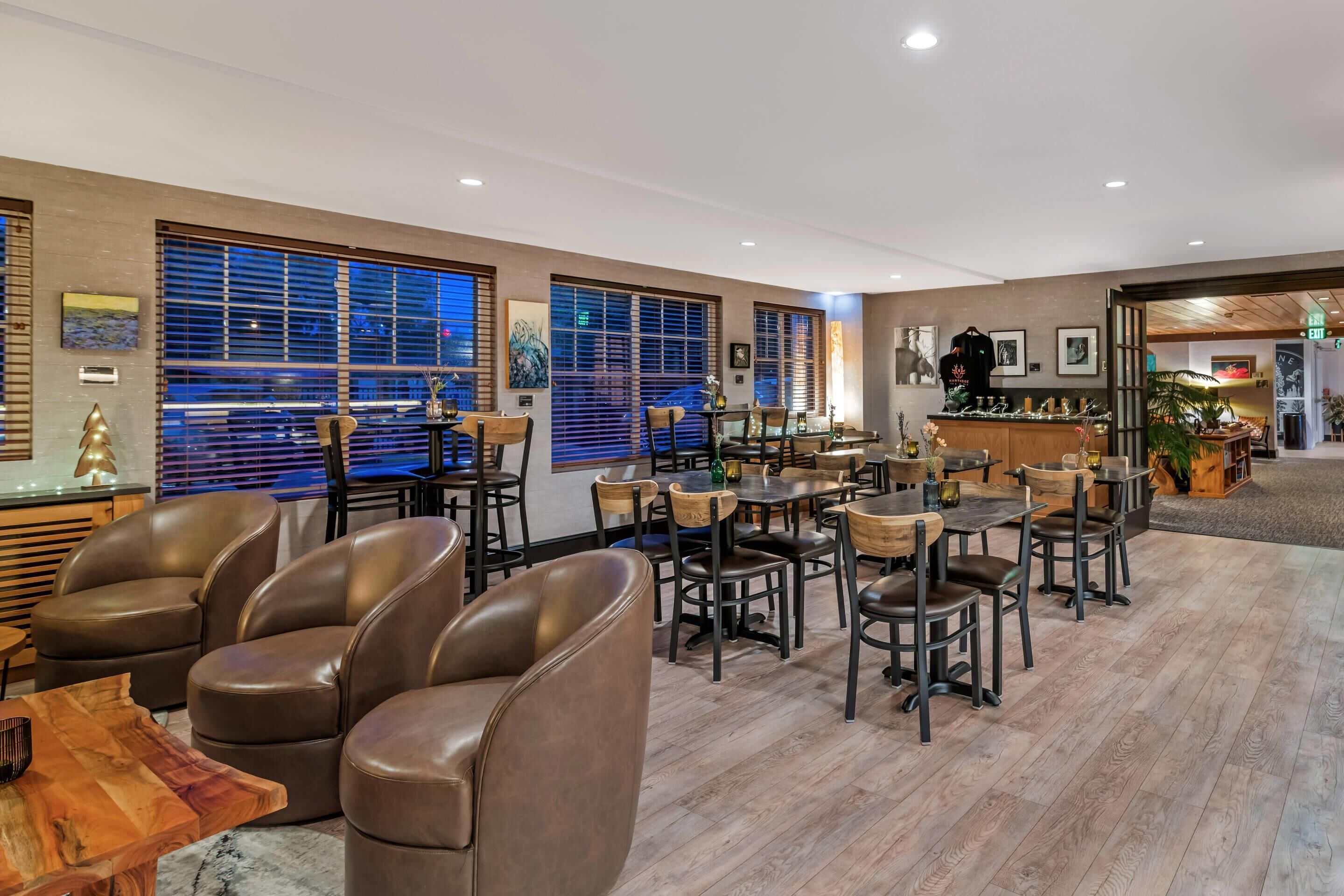 A modern hotel lobby with leather chairs, wooden tables, and large windows with blinds.