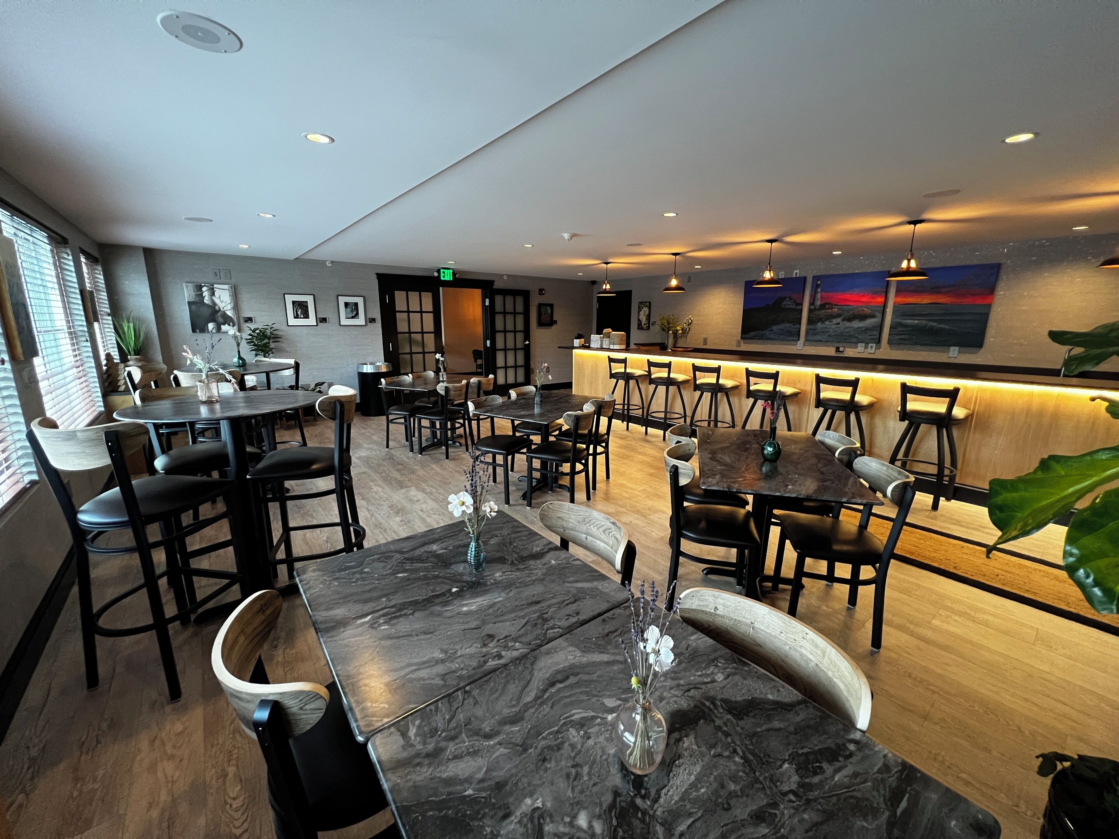 Un restaurant avec des tables en marbre, des chaises noires et un coin bar avec des tabourets.