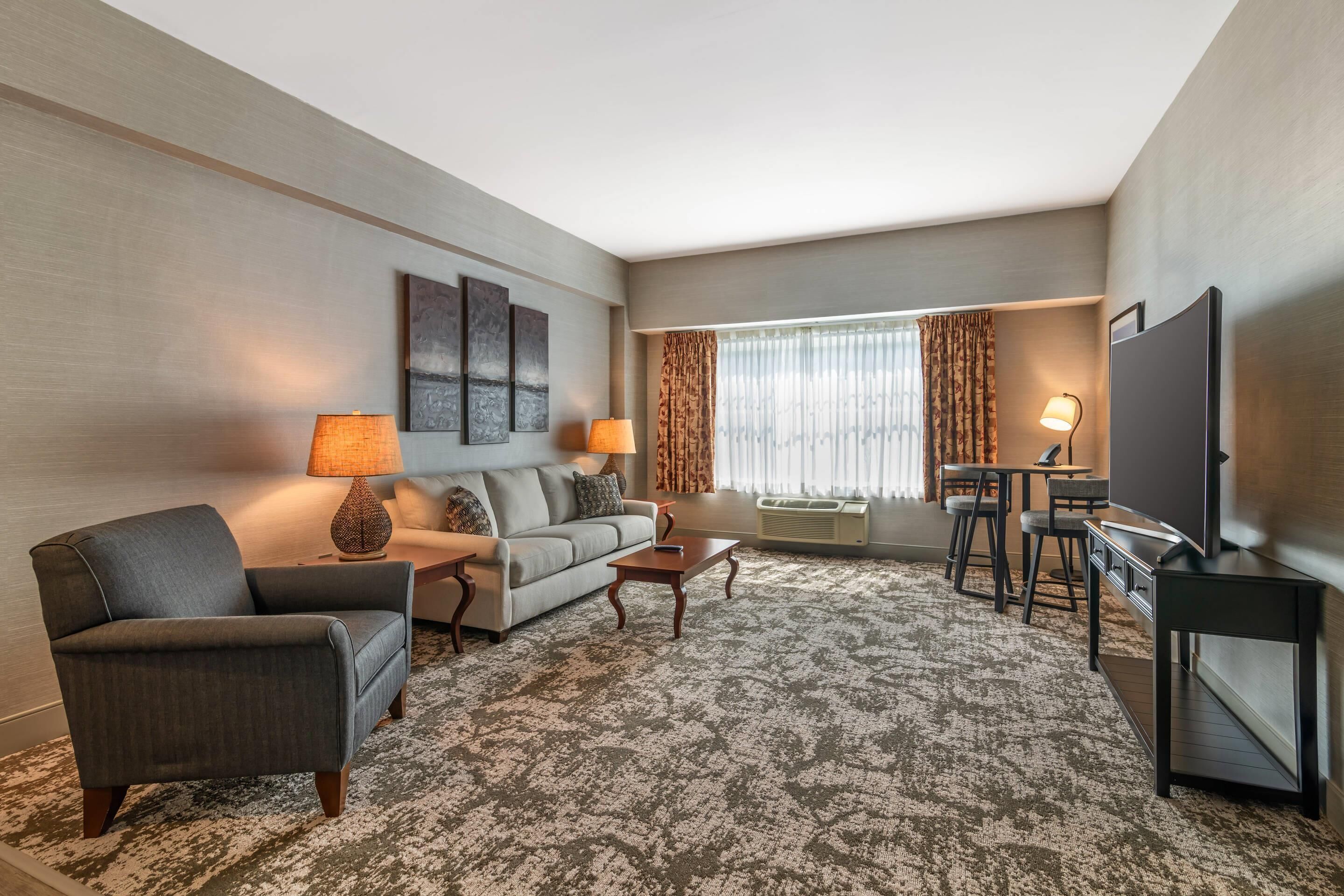 A modern hotel room with a grey sofa, a grey armchair, a coffee table, a TV, and a desk with a chair.