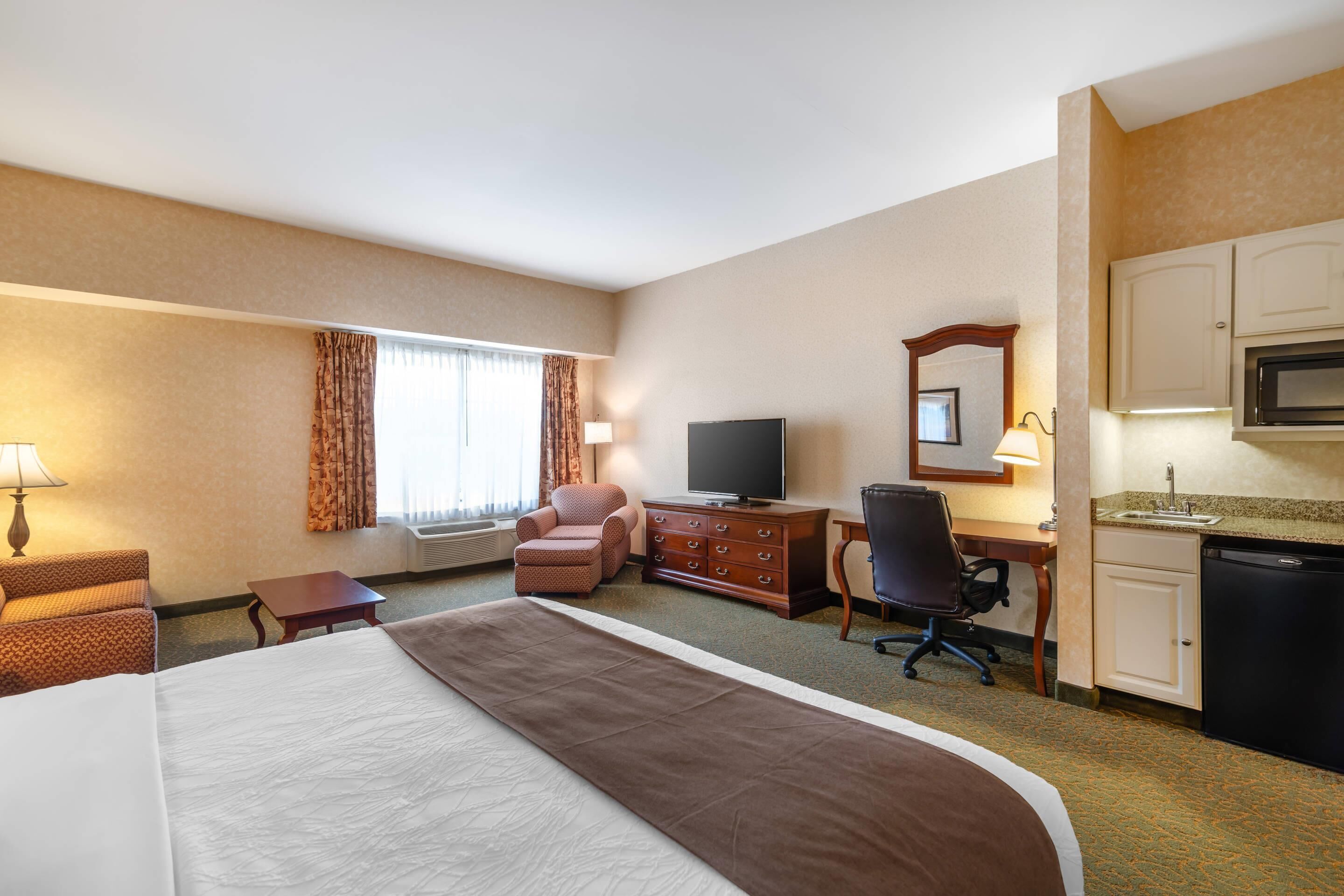 A hotel room with a large bed, a desk with a chair, a television, a small table, a lamp, a window with curtains, and a kitchenette.