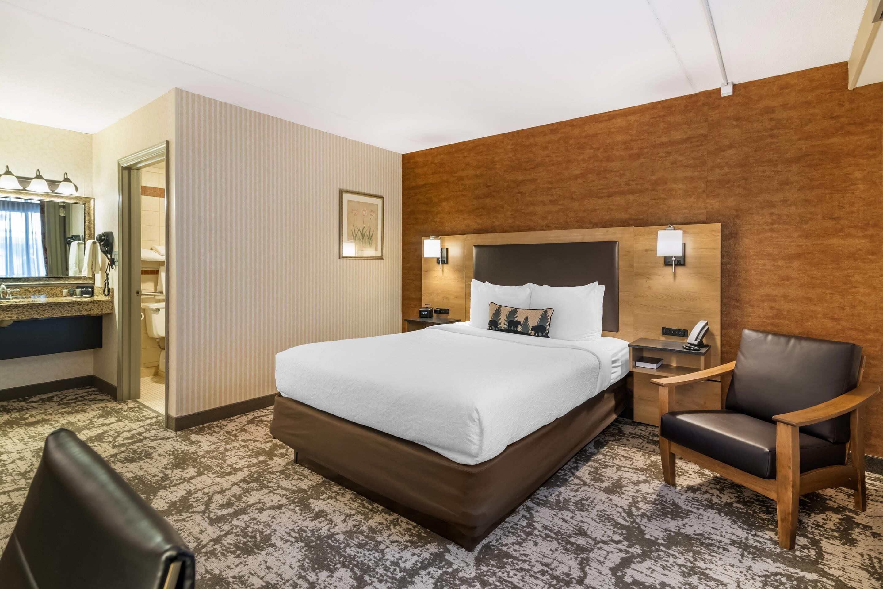 Une chambre d’hôtel avec un grand lit, une table de chevet en bois, une chaise, une salle de bain avec un miroir et une lampe fixée au mur.