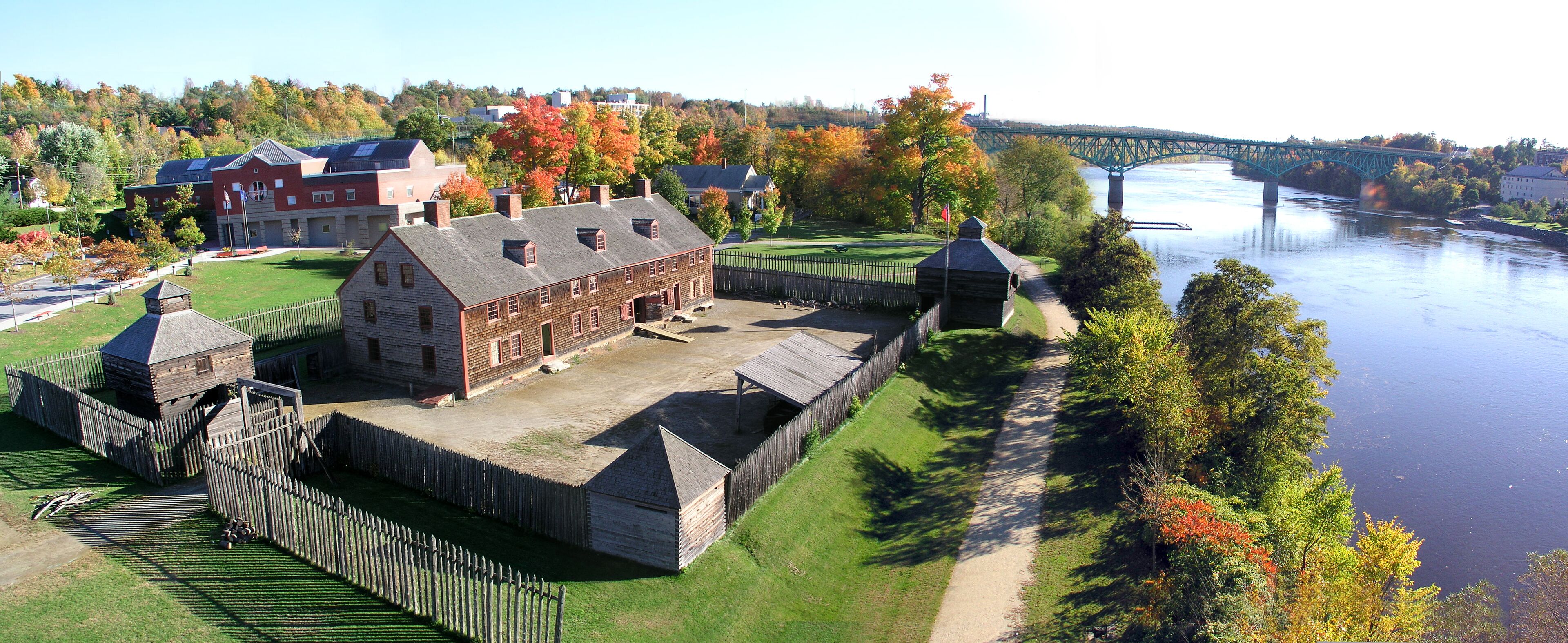 A historic building with a large central structure and smaller outbuildings, surrounded by a wooden fence and a river.