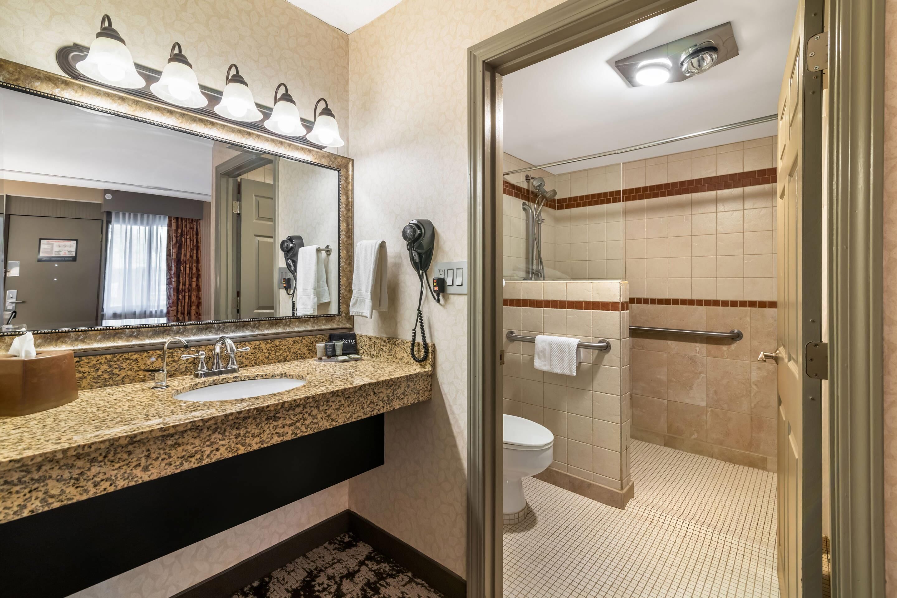 A hotel bathroom with a granite countertop sink, a large mirror, a hairdryer, and a shower area.