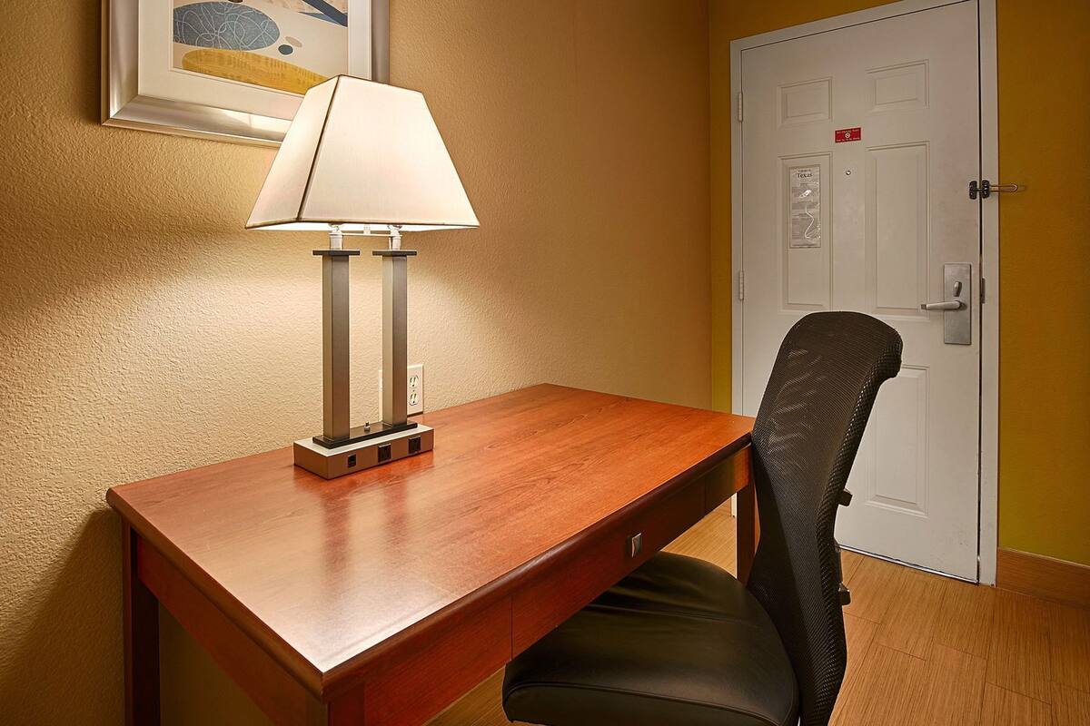 A hotel room with a wooden desk, a chair, a lamp, and a door with a sign.
