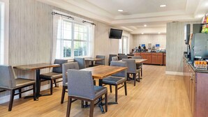 A modern dining area with wooden tables and grey upholstered chairs, a kitchen area with a coffee station, and a flat-screen TV mounted on the wall. - Best Western Cedar Inn & Suites (Angels Camp)