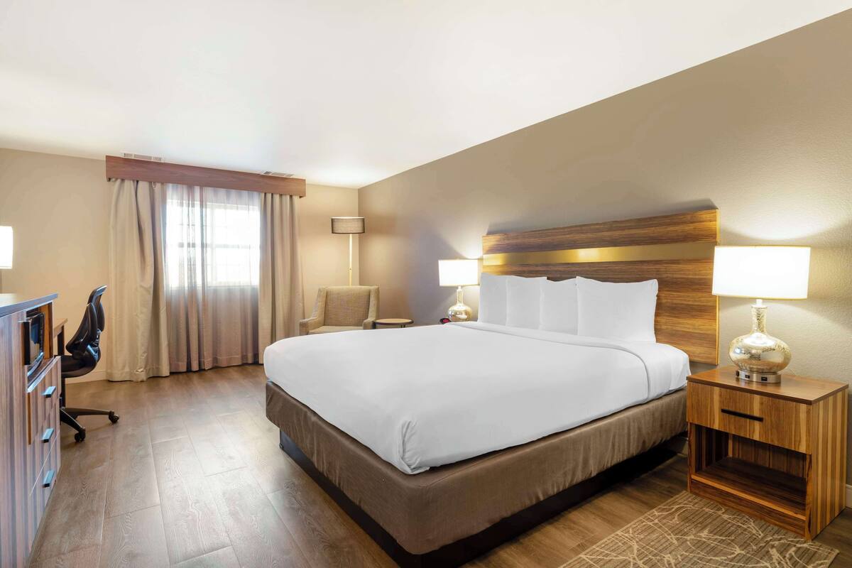 A hotel room with a large bed, two bedside tables with lamps, a desk with a chair, and a window with curtains.