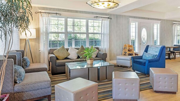 A spacious living room with a sofa, armchairs, ottomans, a glass coffee table, and a large window with curtains. - Best Western Cedar Inn & Suites (Angels Camp)