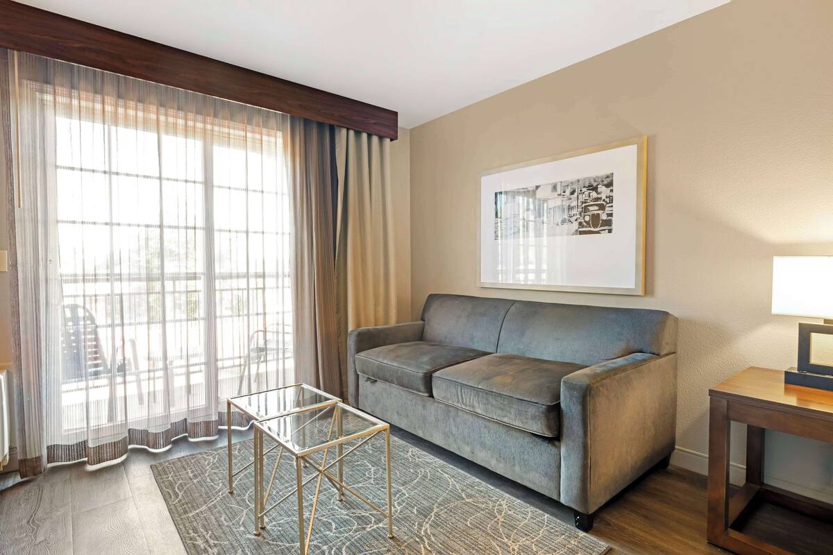 A living room with a grey sofa, a glass coffee table, a wooden side table, a framed picture on the wall, and sheer curtains on the window.