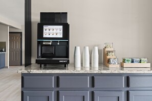 A BREWIT coffee machine with a menu display, stacked white cups, and a coffee bean storage unit on a kitchen counter. - City Express By Marriott Gettysburg (Gettysburg)