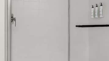 A glass-enclosed shower area with a metal door, a black towel rack, and three bottles of shampoo on the wall.