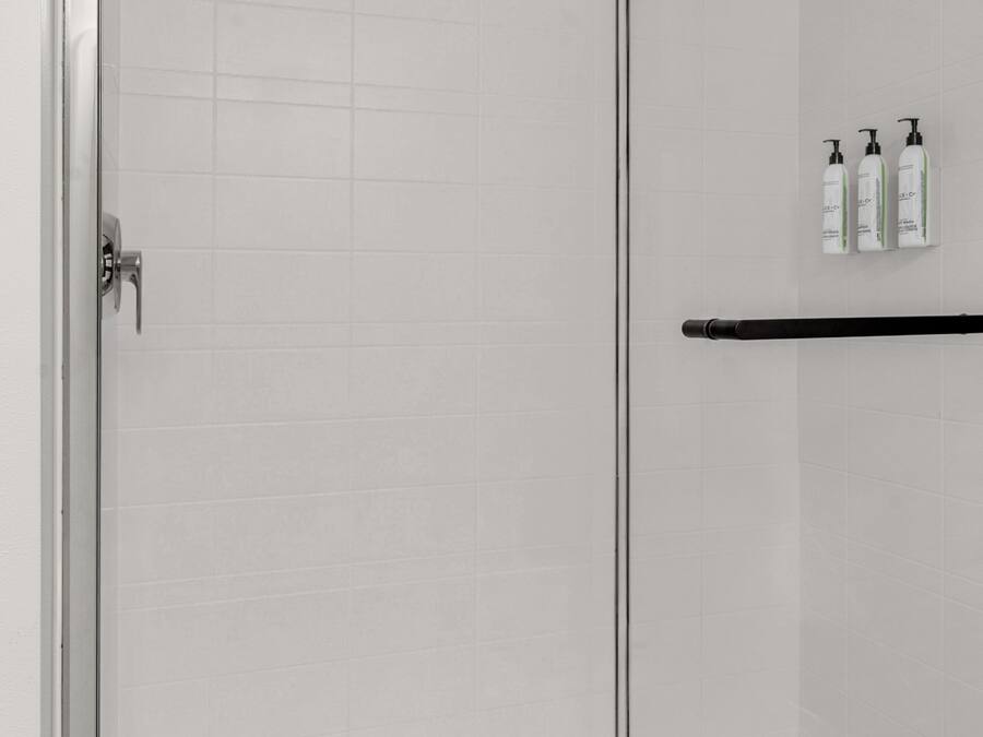 A glass-enclosed shower area with a metal door, a black towel rack, and three bottles of shampoo on the wall.