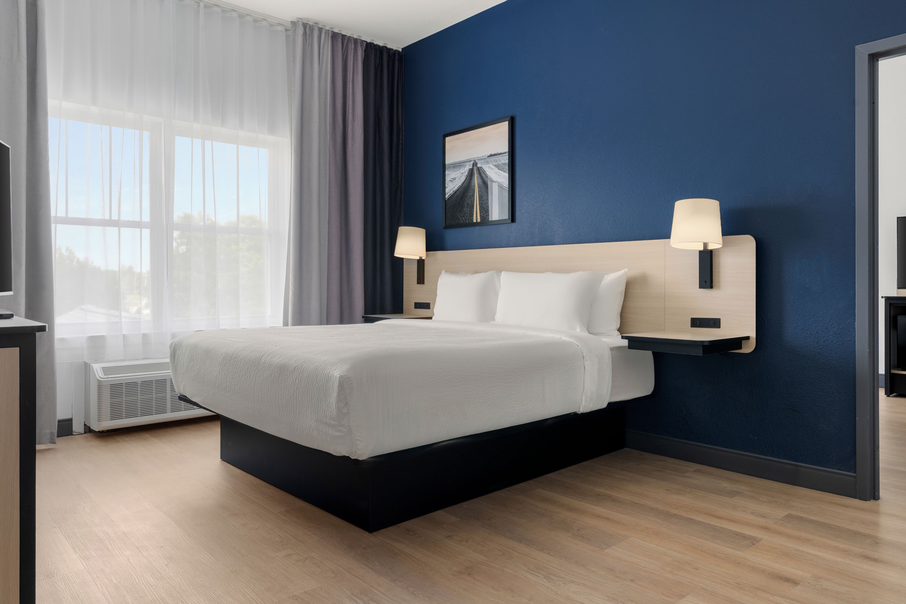 A modern hotel room with a large bed, two bedside lamps, a window with sheer curtains, and a painting on the wall.