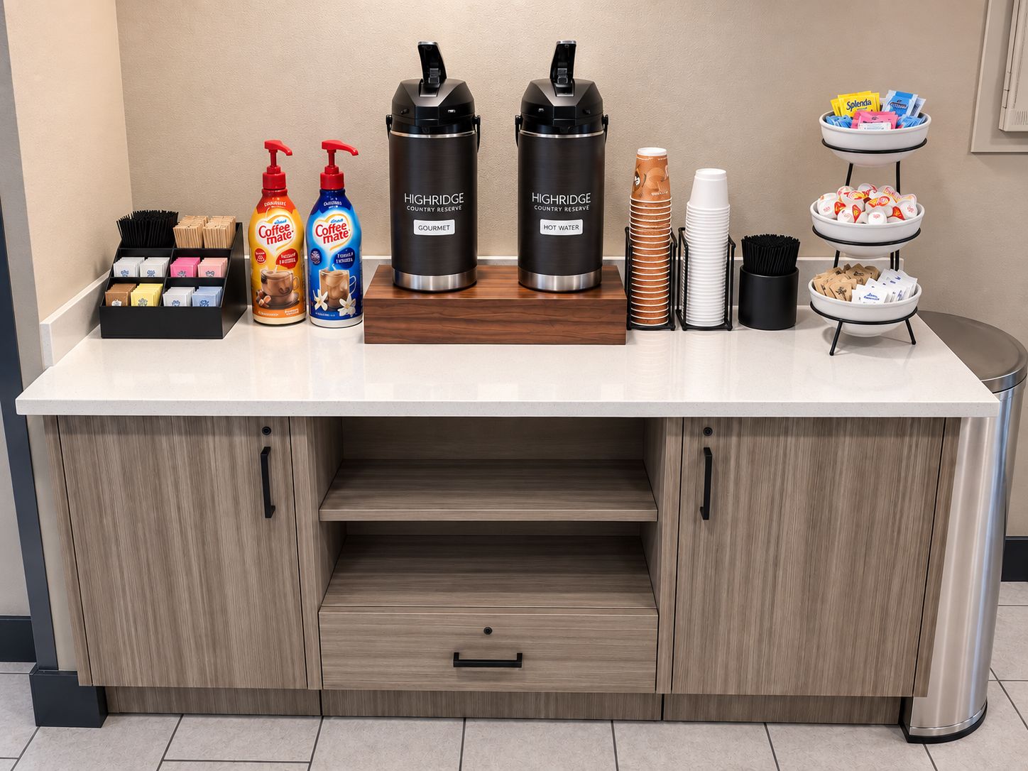 A hotel bar area with coffee dispensers, a hot water dispenser, a tray with snacks, and a coffee station.