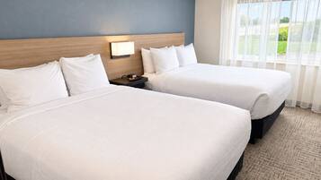 A hotel room with two beds, a nightstand with a lamp, and a view of a green landscape through sheer curtains.