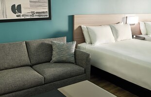 Una habitación de hotel con una cama, un sofá, una mesita de noche, una lámpara y un cuadro en la pared.