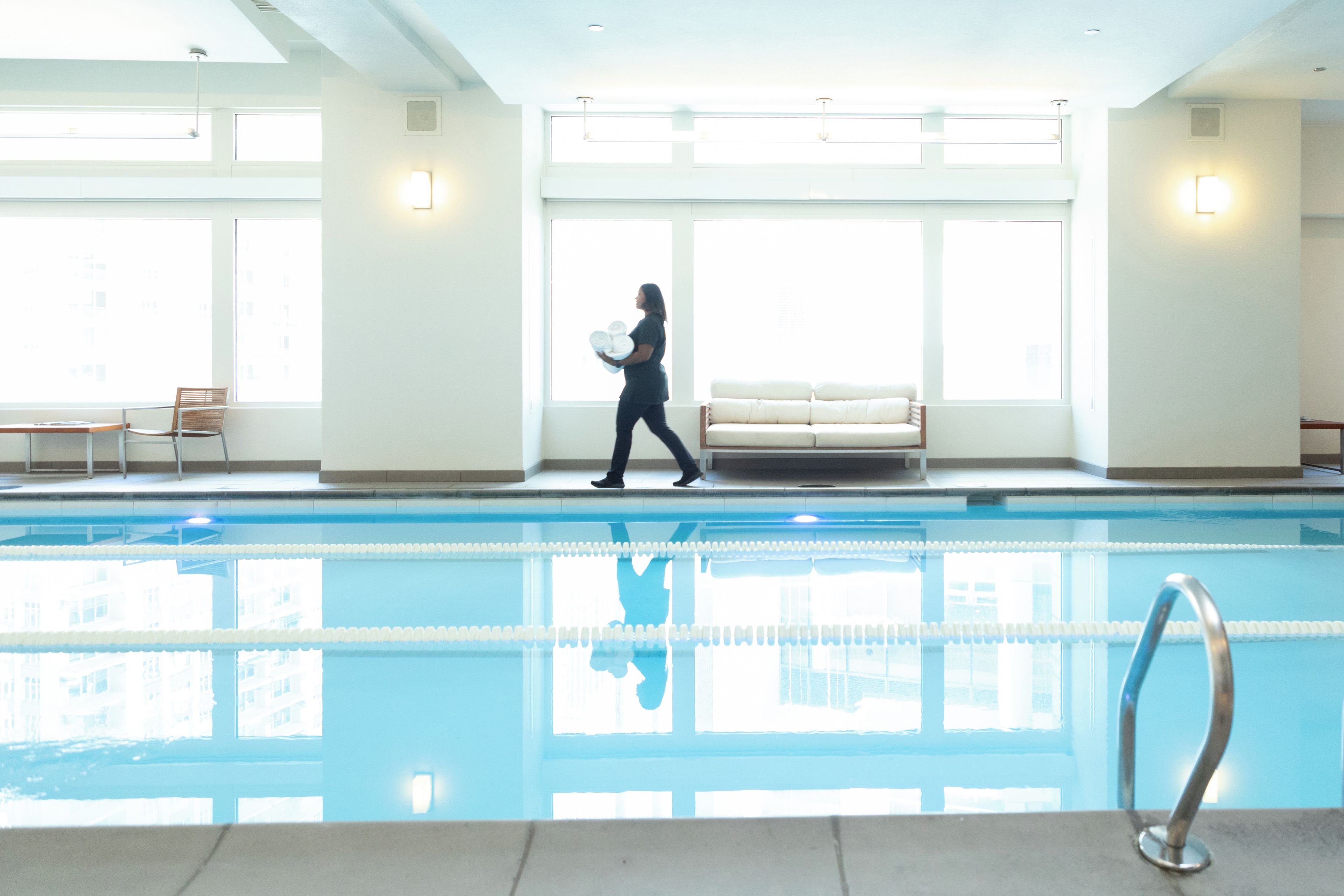 A swimming pool with clear water, a person walking with a towel, and a sofa in a bright indoor space.