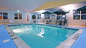 An indoor swimming pool with clear water, surrounded by blue flooring and a poolside area with chairs and umbrellas.