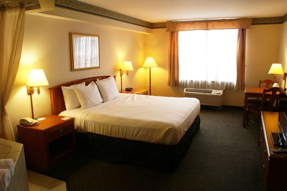 A hotel room with a large bed, two bedside lamps, a desk with a chair, and an air conditioning unit.