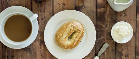 A wooden table with a coffee cup, a plate with a bagel, a glass of orange juice, a jug of milk, and a knife.