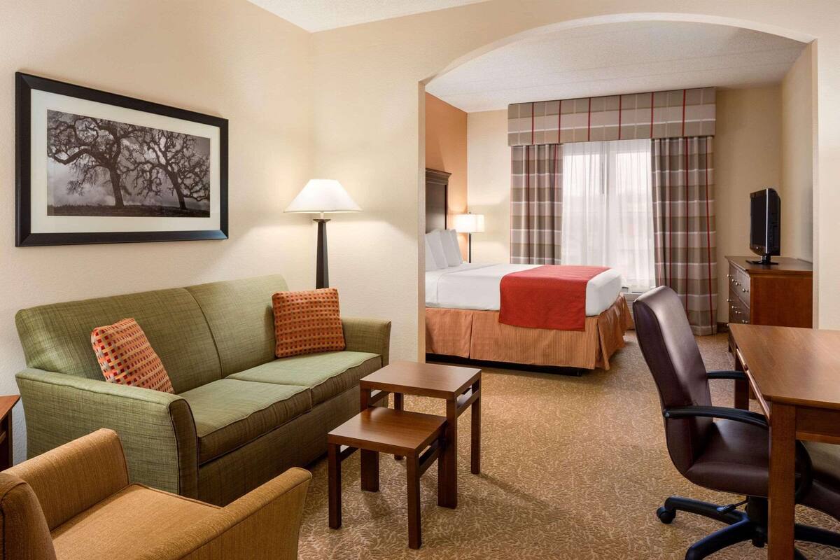 A hotel room with a green sofa, a small table, a brown armchair, a bed with a red blanket, and a television on a wooden stand.