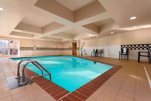 Una piscina interior con un borde de baldosas de diseño ajedrezado, una cubierta de madera y un techo con patrones geométricos.