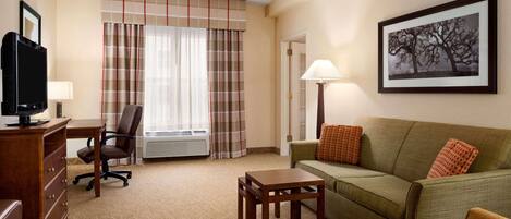 A hotel room with a green sofa, a wooden desk with a chair, a TV, and a framed picture on the wall.