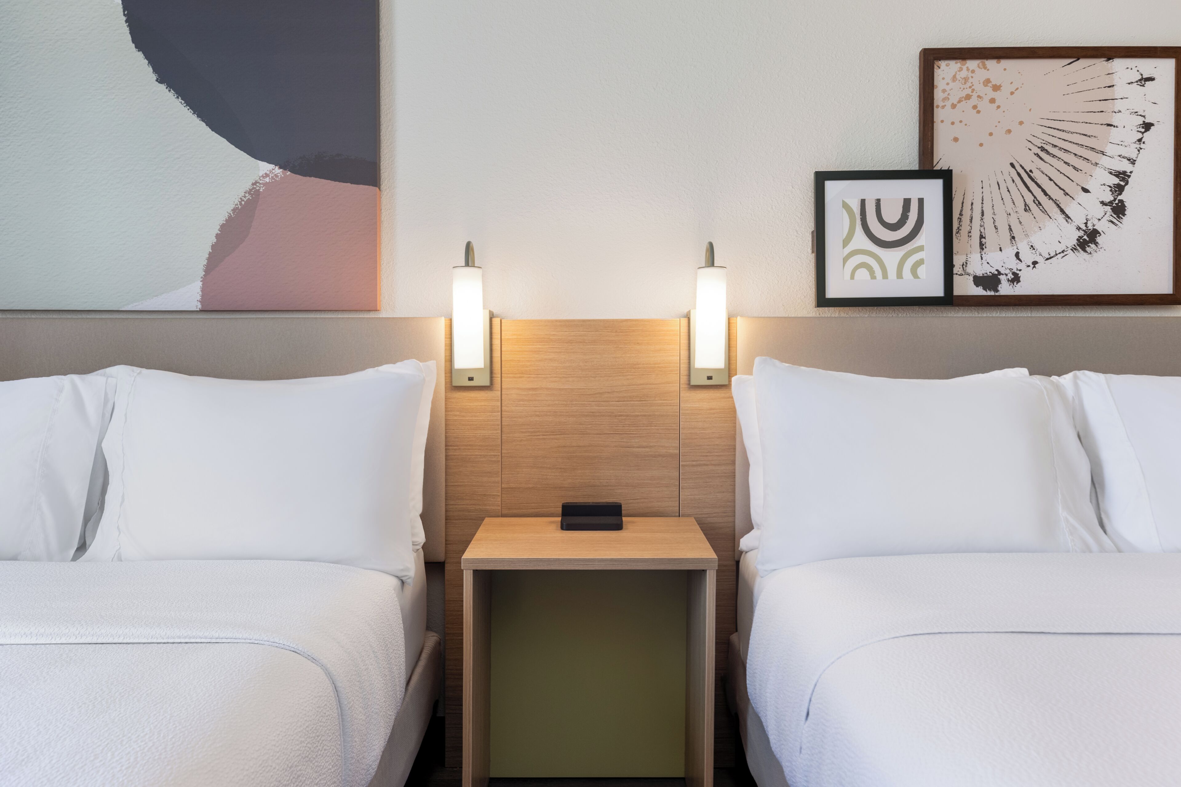 A hotel room with two beds, a nightstand, wall-mounted lights, and artwork.