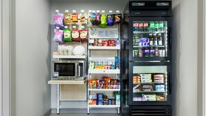A compact kitchenette with a microwave, a small refrigerator stocked with snacks and beverages, and a shelf with various food items. - Spark by Hilton Indianapolis Northeast Fishers (Fishers)