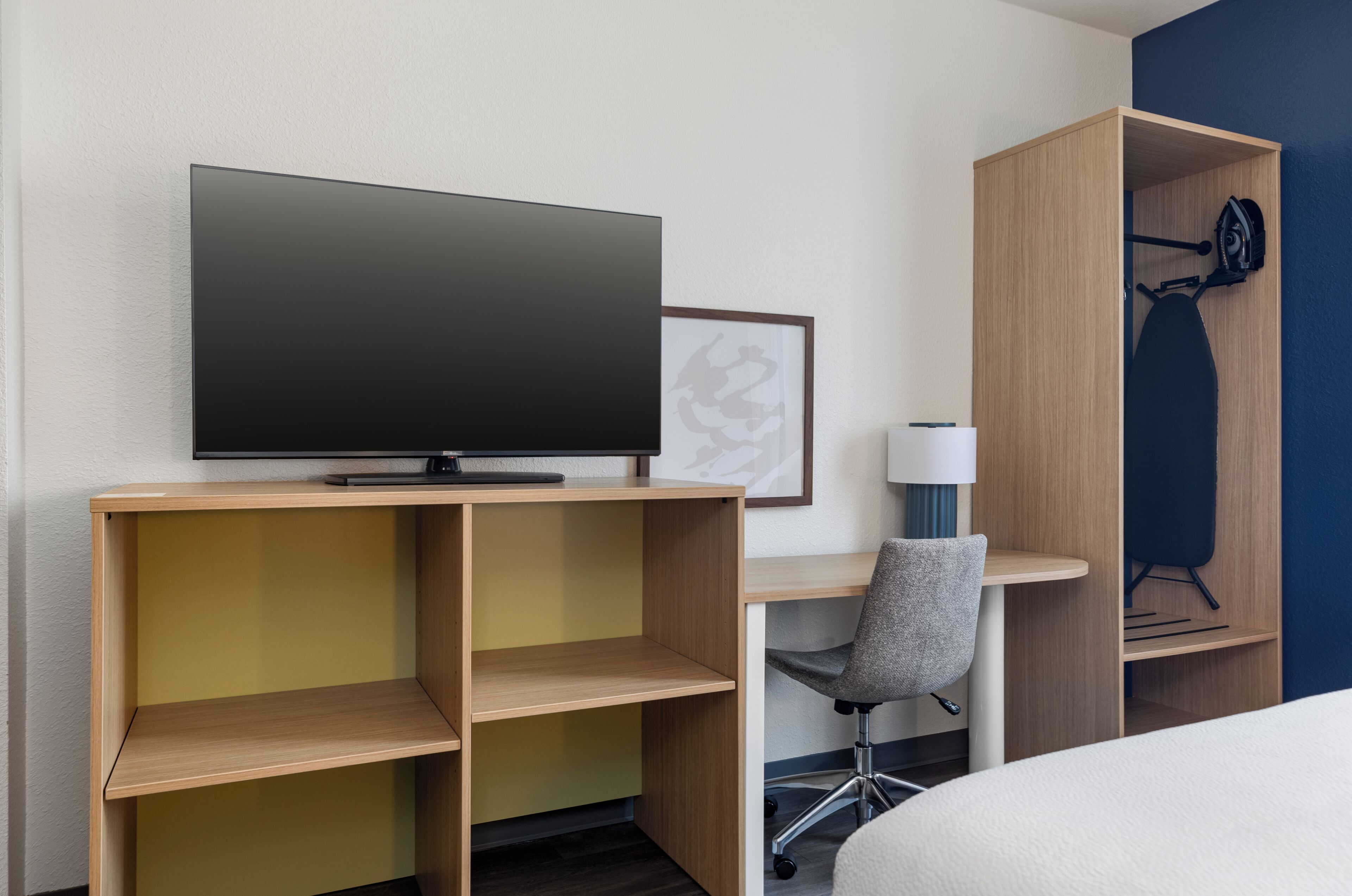 A hotel room with a flat-screen TV, a framed picture, a desk with a chair, and a wardrobe.
