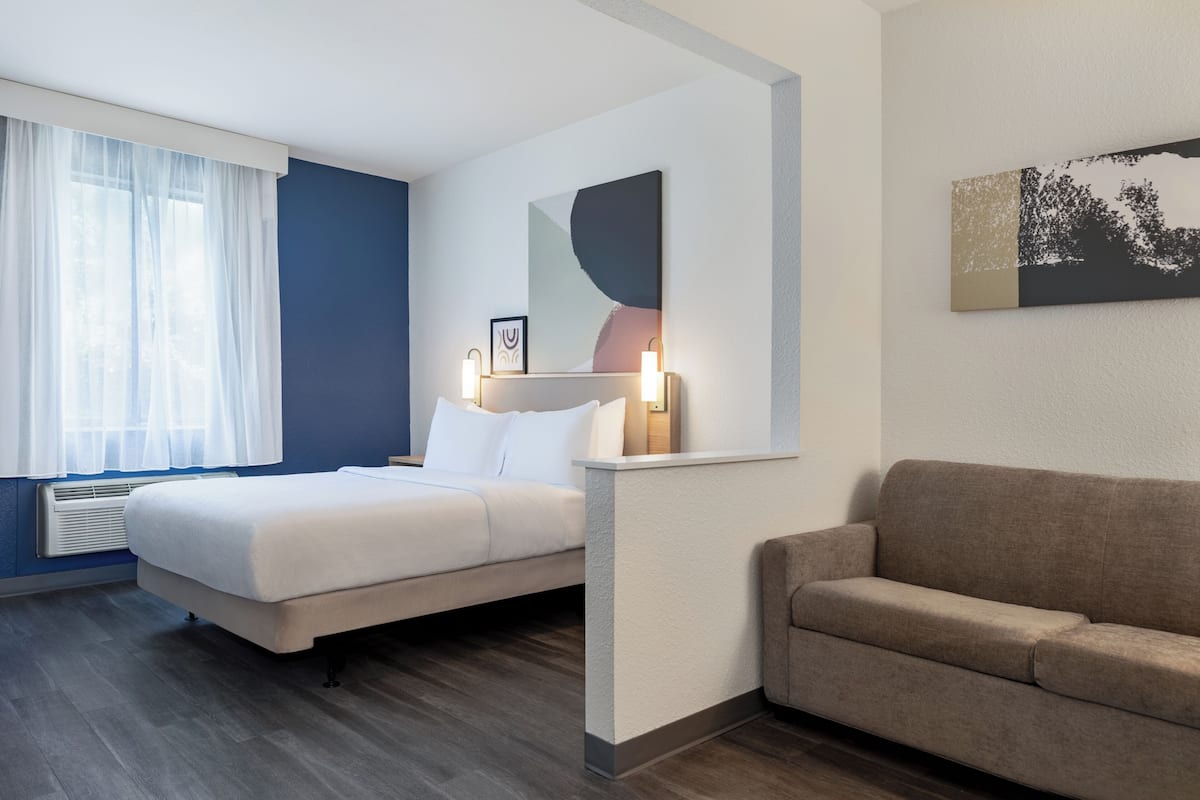 Ein Hotelzimmer mit Bett, Sofa, Fenster mit transparenten Vorhängen, einem Wandbild und einem gerahmten Bild über dem Bett.