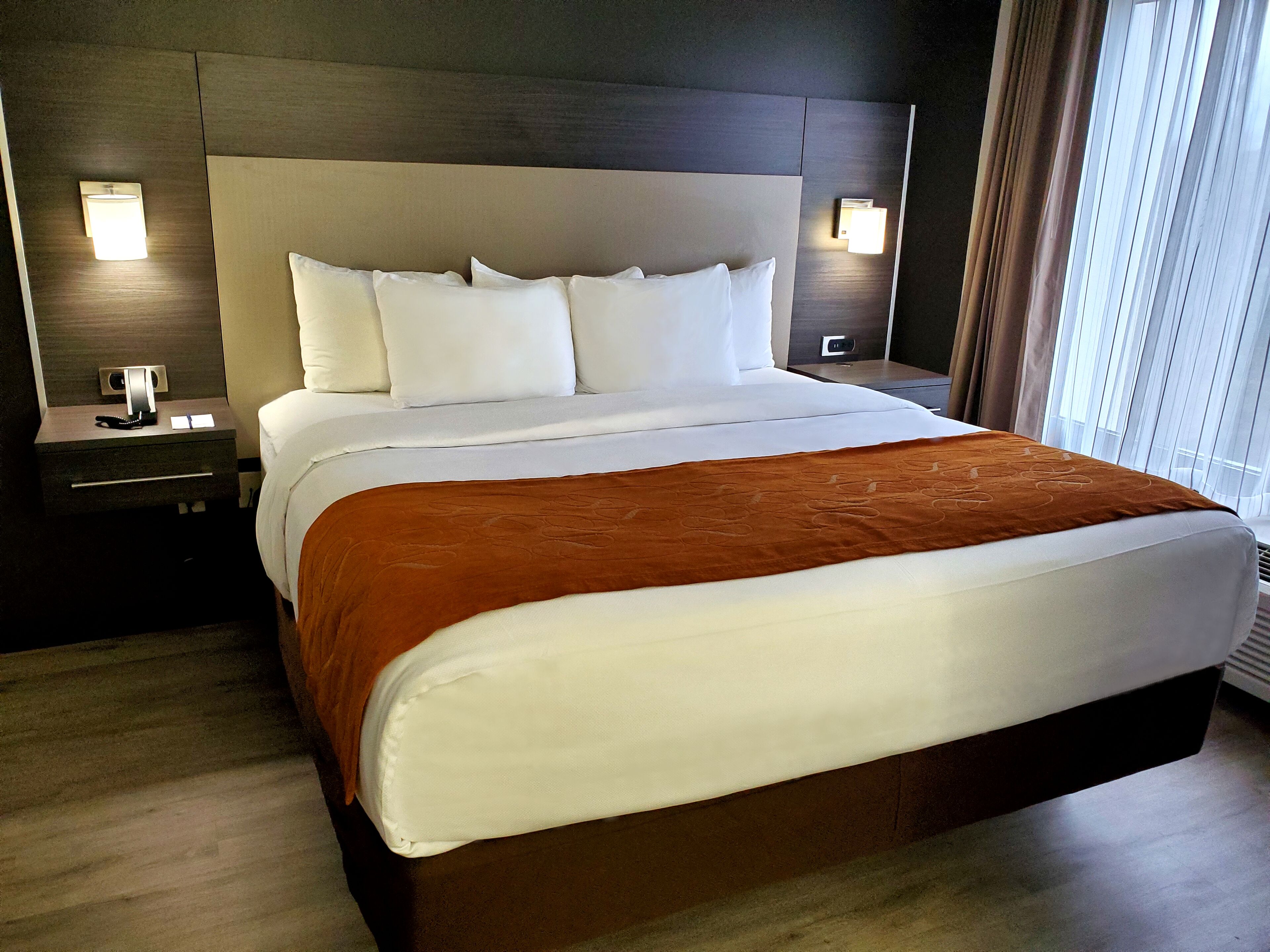 A hotel room with a large bed, a wooden headboard, two bedside tables with lamps, and a window with curtains.