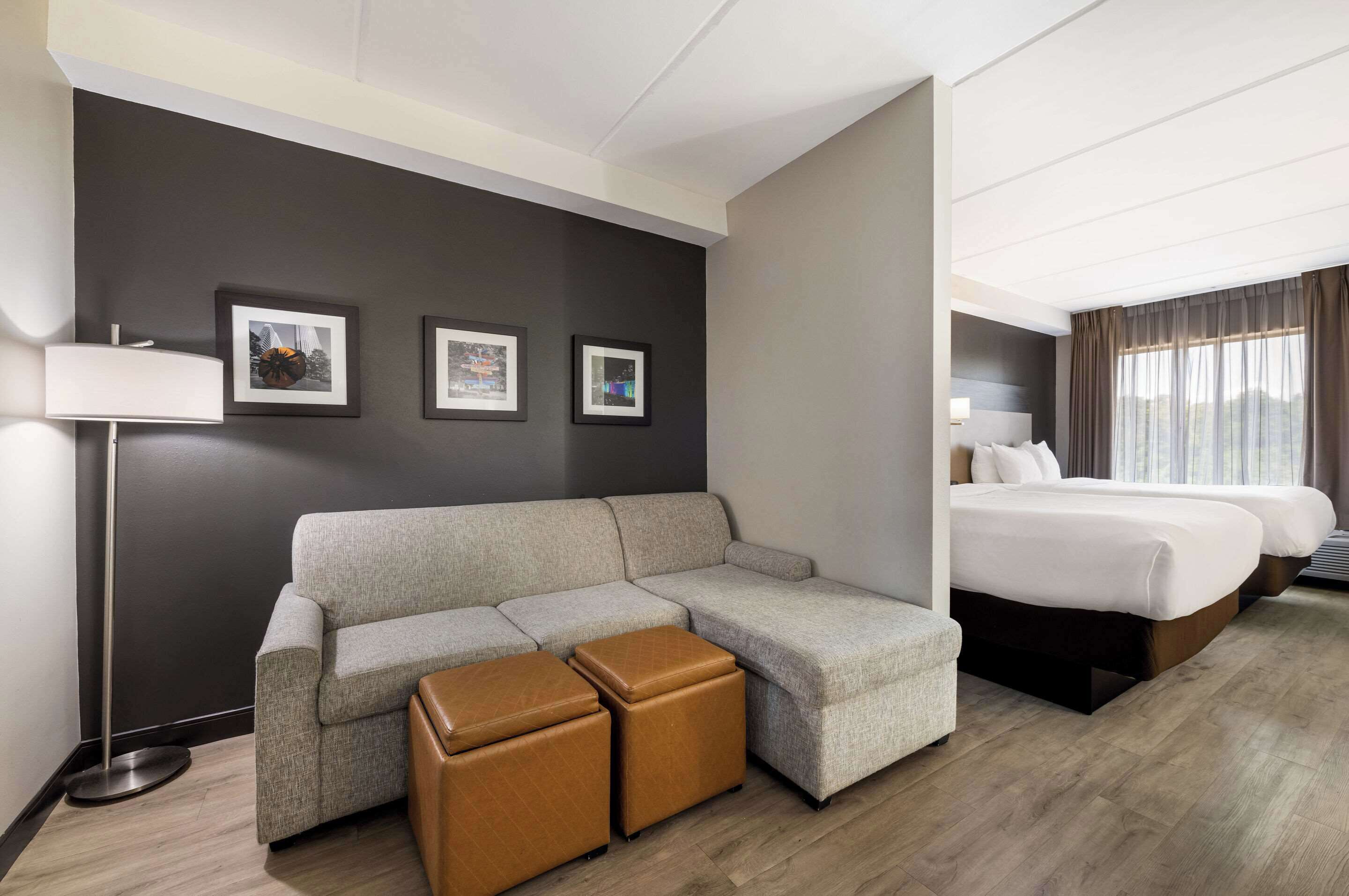 A hotel room with a sectional sofa, two ottomans, a bed with two pillows, a floor lamp, and three framed pictures on the wall.