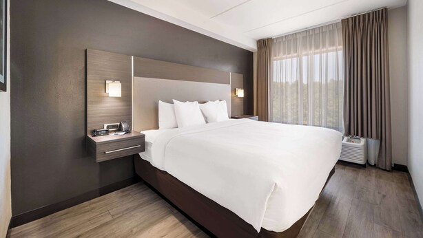 Ein Hotelzimmer mit einem großen Bett, einem Nachttisch mit Lampe, einem Spiegel und einem Fenster mit Vorhängen.