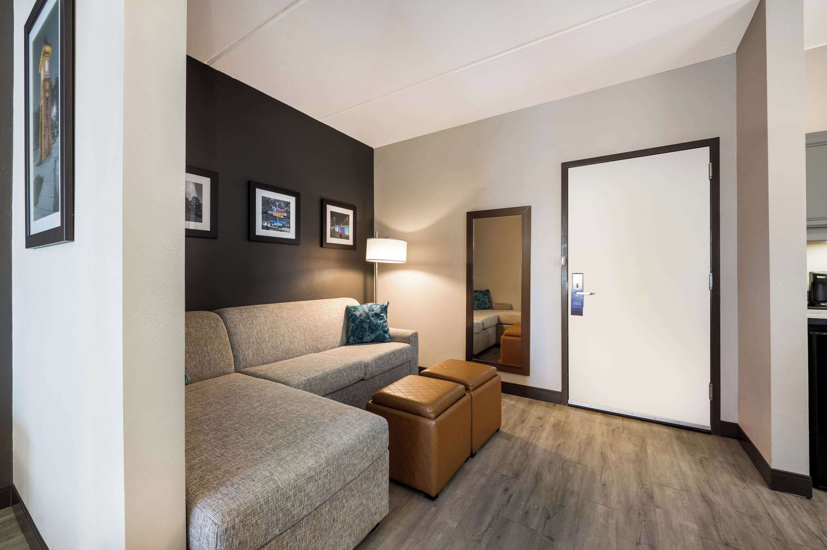 Una habitación de hotel moderna con sofá modular, un pequeño otoman, una lámpara de pie y una puerta con espejo.